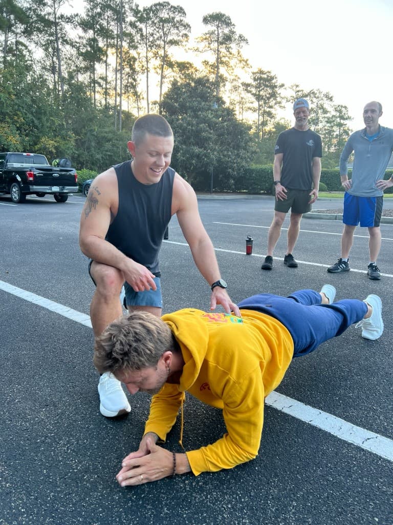 A man steadies another during a forearm plank while others cheer him on